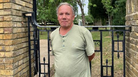 Paul Hayden looks directly at the camera and smiles. He is wearing a green T-shirt and is standing in front of a black gate with a patch of grass behind it. 