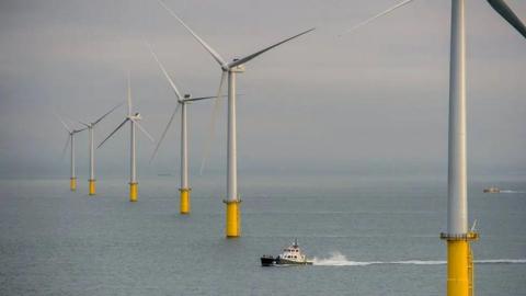 Six white wind turbines with yellow bases in a row in the sea. There is a small boat in between the first two in the image.