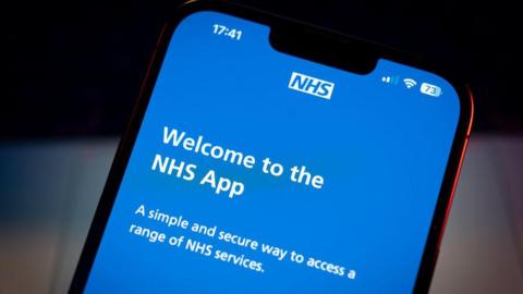 A close-up shot of a phone open on the NHS app. It shows a blue screen with the NHS logo at the top. It reads "Welcome to the NHS App. A simple and secure way to access a range of services".