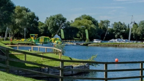 The picture shows Liquid Leisure watersports centre. There is a body of water with grass visible on either side. In the background are green and yellow obstacle courses floating on the water.  