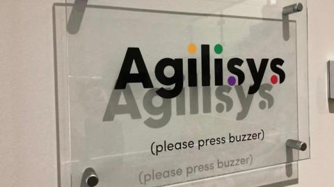 A glass sign mounted on a wall which reads Agilisys (please press buzzer).
