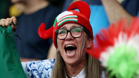 A fan with glasses shouts while hold a flag. She is wearing a red hat with horns and an Italy flag on the front.