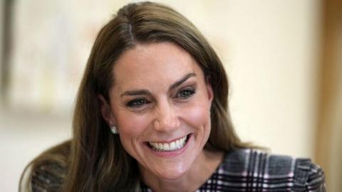 A close up of the face of the Princess of Wales, she is smiling broadly and has long waved brown hair falling over her shoulder and she's wearing a dark grey, black and white tartan print dress. 