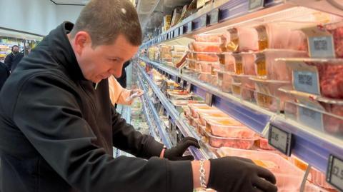 A man stacking supermarket shelves. He wears black gloves and a black sweater, and is reaching into the fresh meat section, picking up a plastic container. Behind him are shoppers and an aisle op