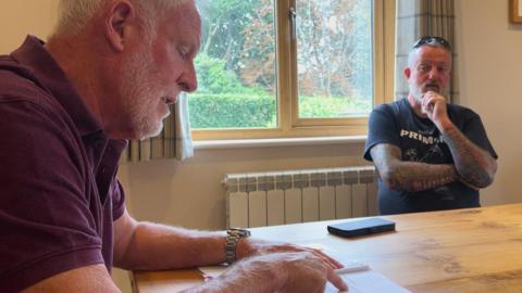 Two men sit at a wooden dining table. There is a document on the table and a phone. There is a window in the background showing greenery and trees.