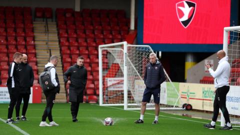 A pitch inspection is conducted at Cheltenham Town's EV Charger Points Stadium with the ball sending water splashing into the air as it hits the ground