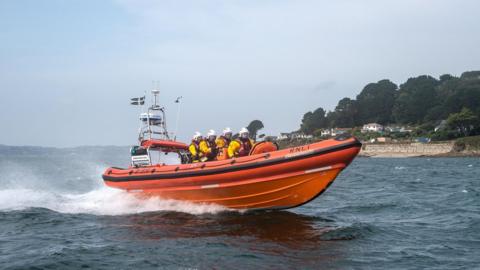 The front view of a orange lifeboat with four people on it. The lifeboat is moving on the water. The crew are wearing yellow life jackets. There is land to the right in the distance with large trees and houses. 