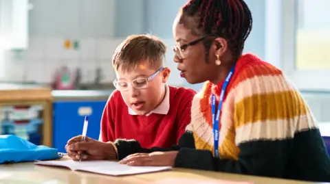 A woman sits with a child in a classroom - she is writing in a workbook with him 