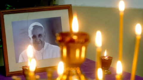 A picture of the Pope in a frame.  It is on a purple table cloth. In front of the picture are multiple candles. 