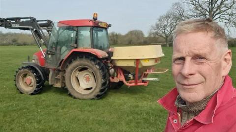 Richard Cornock on his farm in Gloucestershire. He is pictured in a selfie with a red tractor behind him. He is looking at the camera and smiling. 