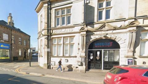 A street corner with a building in the foreground and other smaller buildings in the background.  The main building is constructed from light-colored stone and features ornate architecture. Above the central windows the word MIDLAND is  engraved in capital letters. Below that there is a large arched entrance, above which is an inscription reading 1874. The entrance has black double doors with glass panels and signage for MAVERICK'S. In the right foreground is a red hatchback car.