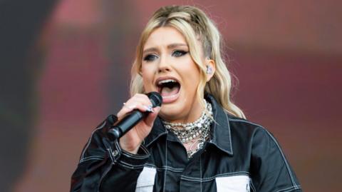 Shot of Ella Henderson performing on stage. She has long blonde hair and is wearing a black jacket and elaborate necklace. She is holding a microphone in her right hand.