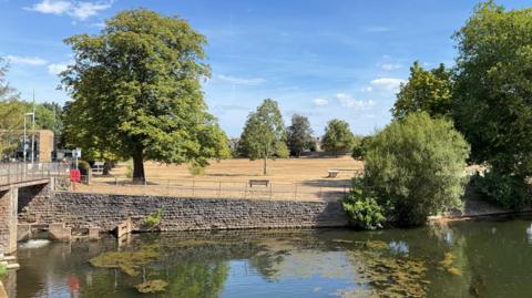 View of a park by a weir with brown grass and green trees.