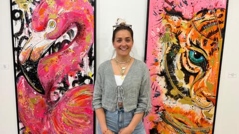 Woman smiling with brown hair standing in front of two brightly coloured paintings on a white wall