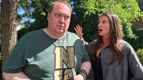 A man holding an award looking scared, while a woman gives him a scary look with her arm up like a claw