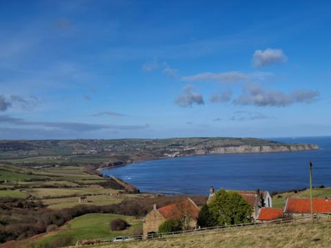 A bird's eye view looking out over corner of a coastline of countryside and sea with fields and houses in distance under a blue sky