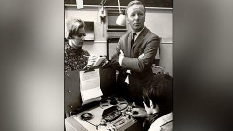 A sharply dressed man in a suit stands with his arms folded whilst a woman takes notes onto a a pad of paper. Another women sits in-front of an old fashioned tape reel player