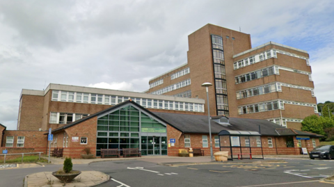 A large brick built hospital with a shelter in front. In front is a large single-storey building with a triangular glass-fronted entrance. Behind it is a three-storey building with long, uninterrupted windows along each floor. To the right is a seven-storey building in the same style, with a large stairwell structure just off-centre. The buildings are arranged in a T-shape with the tallest building at right angles to the other two.
