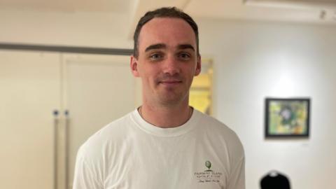 A man with short brown hair, looking at the camera. He is wearing a white t-shirt. There is a white wall and white door in the background. 