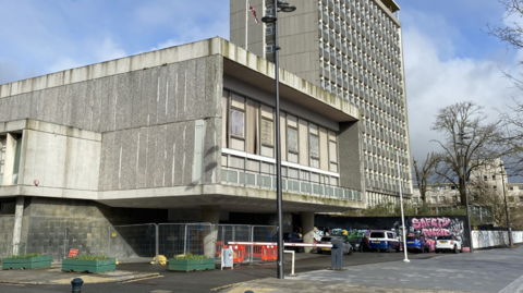 A 1960s style office building with a multi-storey building in the background and some graffiti on a wall and some metal fencing around part of it.