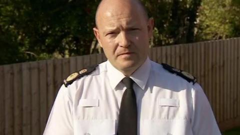 Chief Constable Craig Guildford looking forward towards the camera. He is wearing a white shirt with a black tie with black and gold epaulets on his shoulders. 