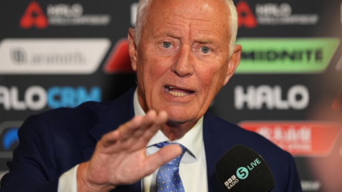 Barry Hearn holds his right hand out in front of him as he talks in a media conference, wearing a mostly light blue tie, white shirt and blue smart jacket