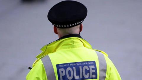 A Greater Manchester Police officer pictured from behind wearing a high visibility jacket, and police hat