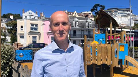Deputy Jonathan Renouf is standing in front of a play park, smiling and wearing a blue shirt.