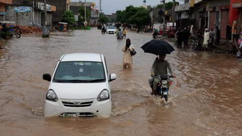 A car, motorbike and people walking - all partially submerged by flood waters on a street.