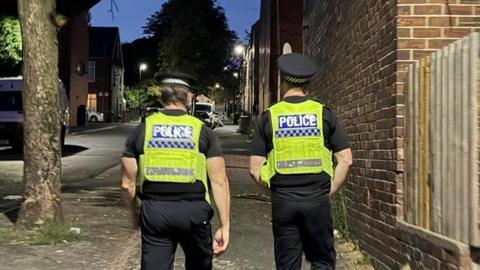Two police officers walking through the Page Hall area of Sheffield. They are wearing high vis jackets and are walking on a road illuminated by streetlights.