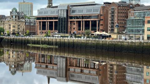 A large imposing court building made from smooth red stone with large dark windows reflected in a river running along its front.