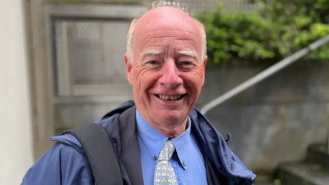 A man wearing a blue shirt with a tie and a dark blue jacket on top, smiling at the camera. The background of stairs and a door is blurred.