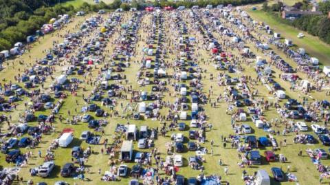 An aerial photo of a car boot sale in a large field. More than one hundred cars are parked in neat lines with goods being sold in front of them, with lots of people milling around