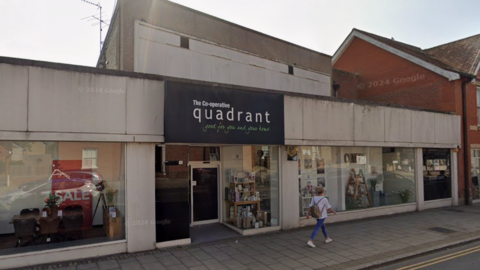 The front of the Quadrant department store in Braintree. It is a single-storey concrete block, with a woman in white and blue jeans walking past the store. It is a sunny day.