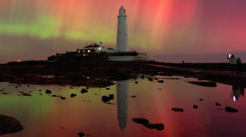 A white lighthouse with a blaze of red and green lights in the sky behind it, all reflected in a pool of water in front