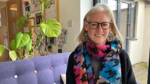Catherine Chapman sitting on a purple chair, smiling at the camera. She is wearing a bright scarf and glasses.