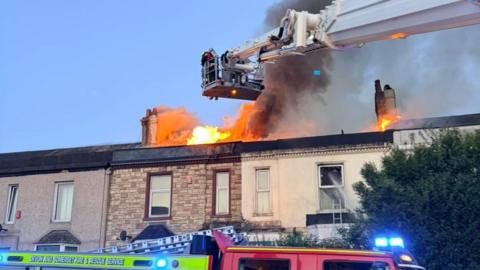 The photo shows a serious house fire affecting the roof of a terrace of homes. Large flames are visible along the roofline and thick black smoke is billowing into the sky while firefighters use an aerial ladder platform to tackle the blaze from above. In the foreground, a red fire engine is parked on the street with emergency lights on and equipment visible in an open side compartment.