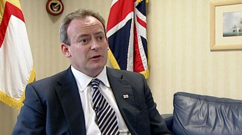 Deputy Lyndon Trott, a man with a black suit and a black and white metallic tie sitting in front of a Guernsey and a Great British flag. The man is wearing a Guernsey flag on his lapel. He has greying hair and brown eyes. 