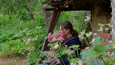 A woman sniffs a bunch of flowers surrounded by green garden foliage.