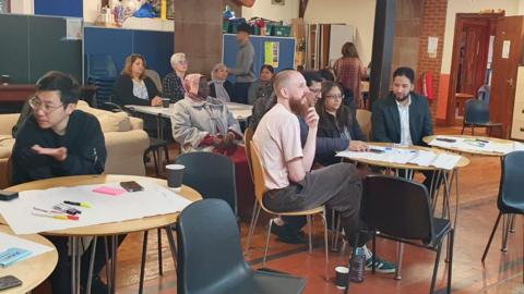 About 15 people sit around tables with pens and paper in a community hall. The tables are round and wooden with large sheets of white paper on them and colourful pens. People are talking to each other, and others can be seen walking around in the background.