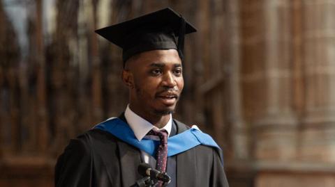 Aaron Agbanu speaks in front of microphone on a lectern. He wears a black graduation hat and robe with a blue caped strap across his tie. He is inside the grand interior of Chester Cathedral.