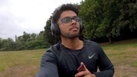 Mridul is running in a field, with his phone in hand and headphones on his head, while wearing glasses and a gold necklace