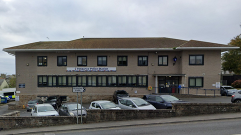 A large, two-storey, detached building with a banner which reads Penzance Police Station. There are cars parked in front of it and a couple of police cars are parked behind it to the left. There is a road in the foreground.
