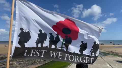 A flag saying 'lest we forget' flying in the forefront, with a beach behind and an man in military garb in a wheelchair