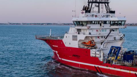 A red and white research-style vessel on a blue sea. The boat is equipped with cranes and surveying technology. The coast can be seen in the distance. 