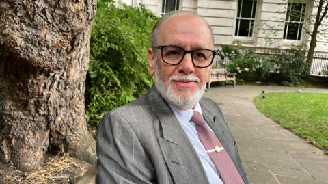 George Psaradakis in a mauve tie, grey suit and black glasses sits on a bench in a park with a tree trunk behind him.