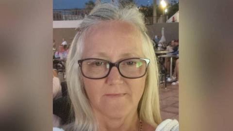 Angela is a blonde lady. She is taking a picture of herself. 
She is wearing rectangular glasses, a white top and a good chain. 