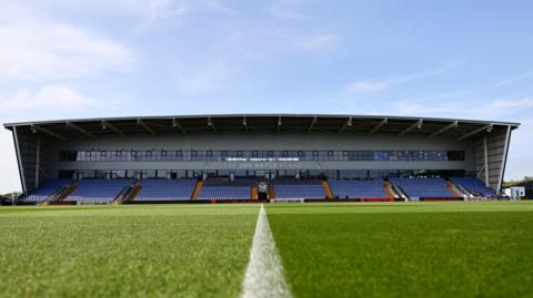 A ground-level view of the Joe Royle Stand at Oldham's Boundary Park on a sunlit early evening.