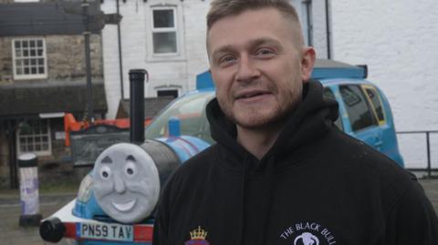 Alex Birkinshaw, a man with short hair and a stubble beard, standing in front of a - peaking from behind him - with the face of Thomas the Tank Engine constructed on its bonnet. The car has been sprayed blue with red stripes. Thomas's smiling face is stuck to the front with a funnel poking out the top and imitation wheels have been stuck to the sides of the car to complete the look.