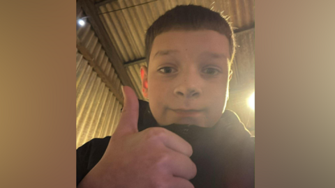 Aaron Anderson is wearing a zipped-up coat and is holding a thumbs up next to the camera. He is taking a selfie and smiling.